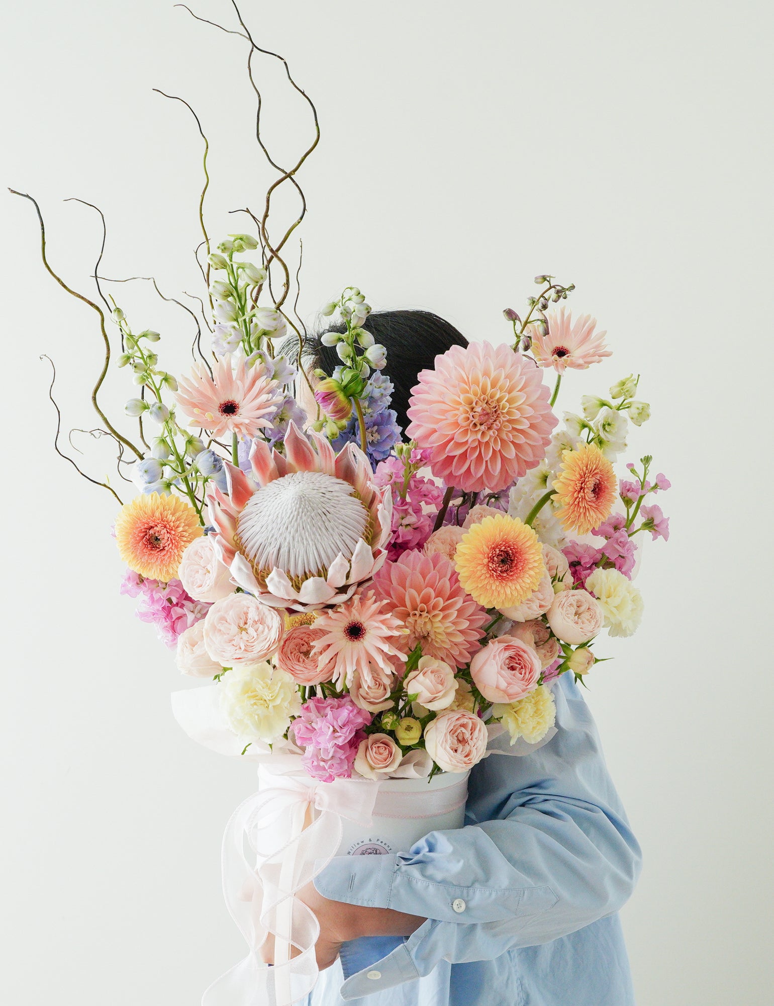 A person holding a luxurious flower hat box with a variety of blooms including protea, peonies, and willows, partially obscuring their face.