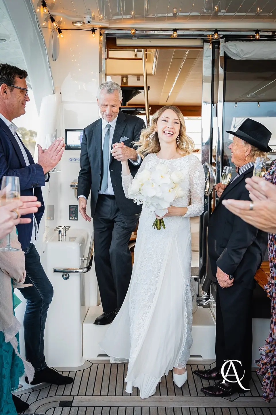 Bride stepping out of a yacht cabin holding a bouquet of white garden roses and phalaenopsis orchids as guests applaud