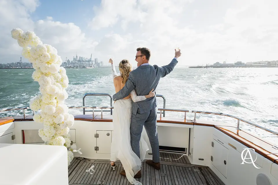 Couple celebrating at the stern of a yacht beside a white hydrangea column, with the Auckland skyline behind them