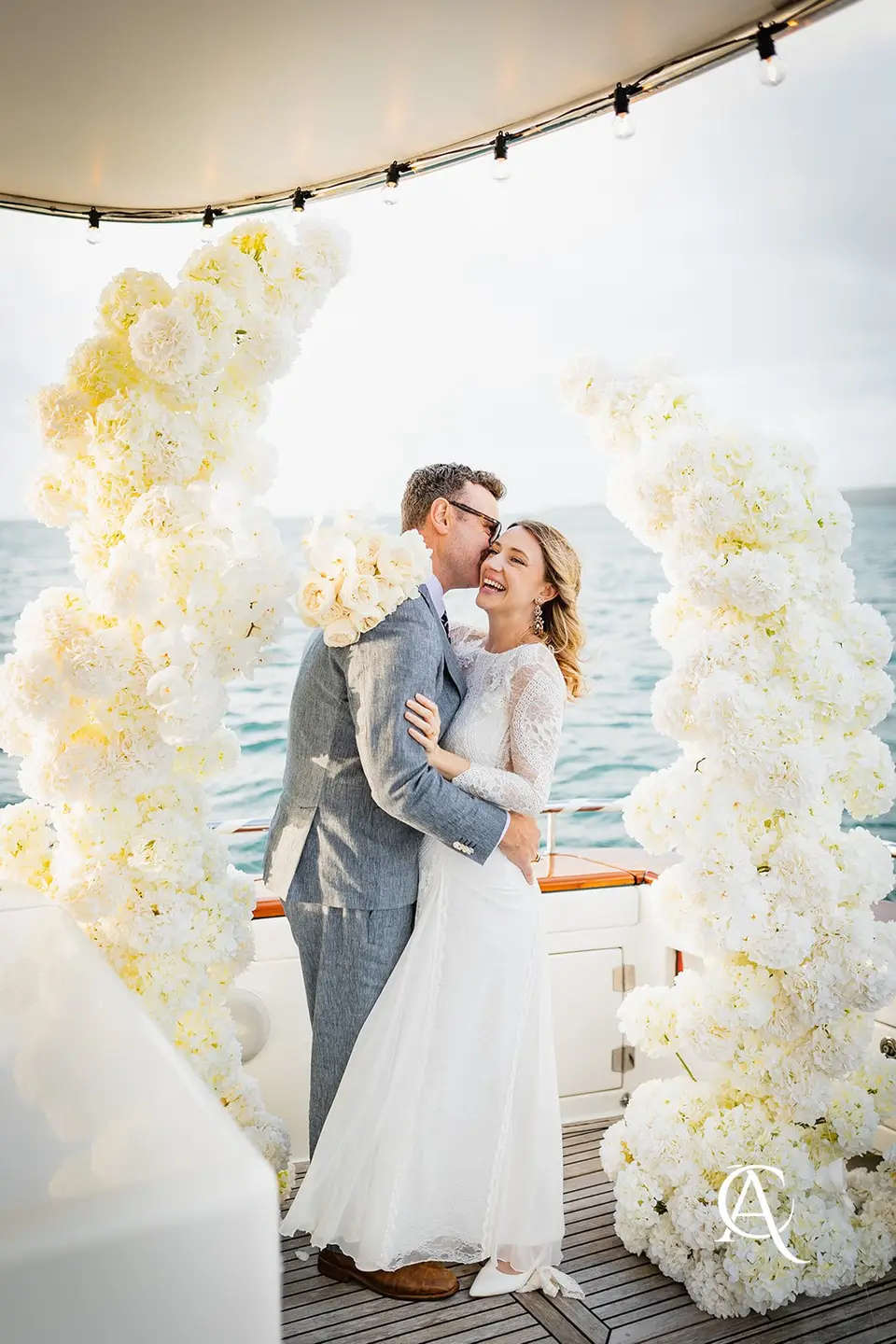 Couple laughing between two twisting white hydrangea columns under a yacht canopy strung with festoon lights