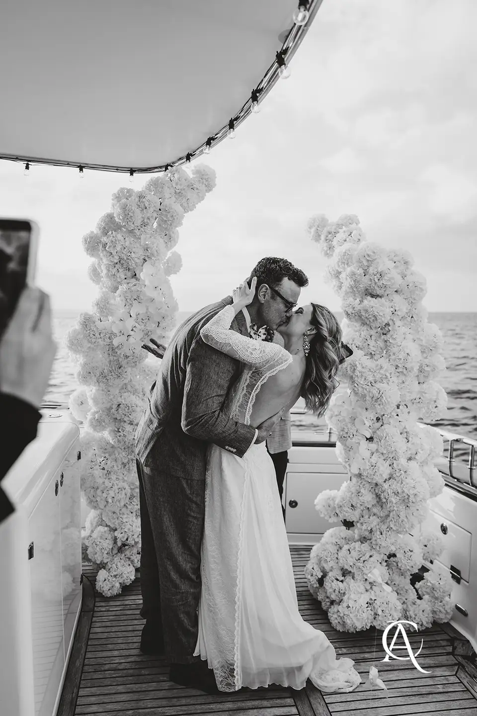 Black and white photo of a couple kissing between two hydrangea columns under a yacht canopy
