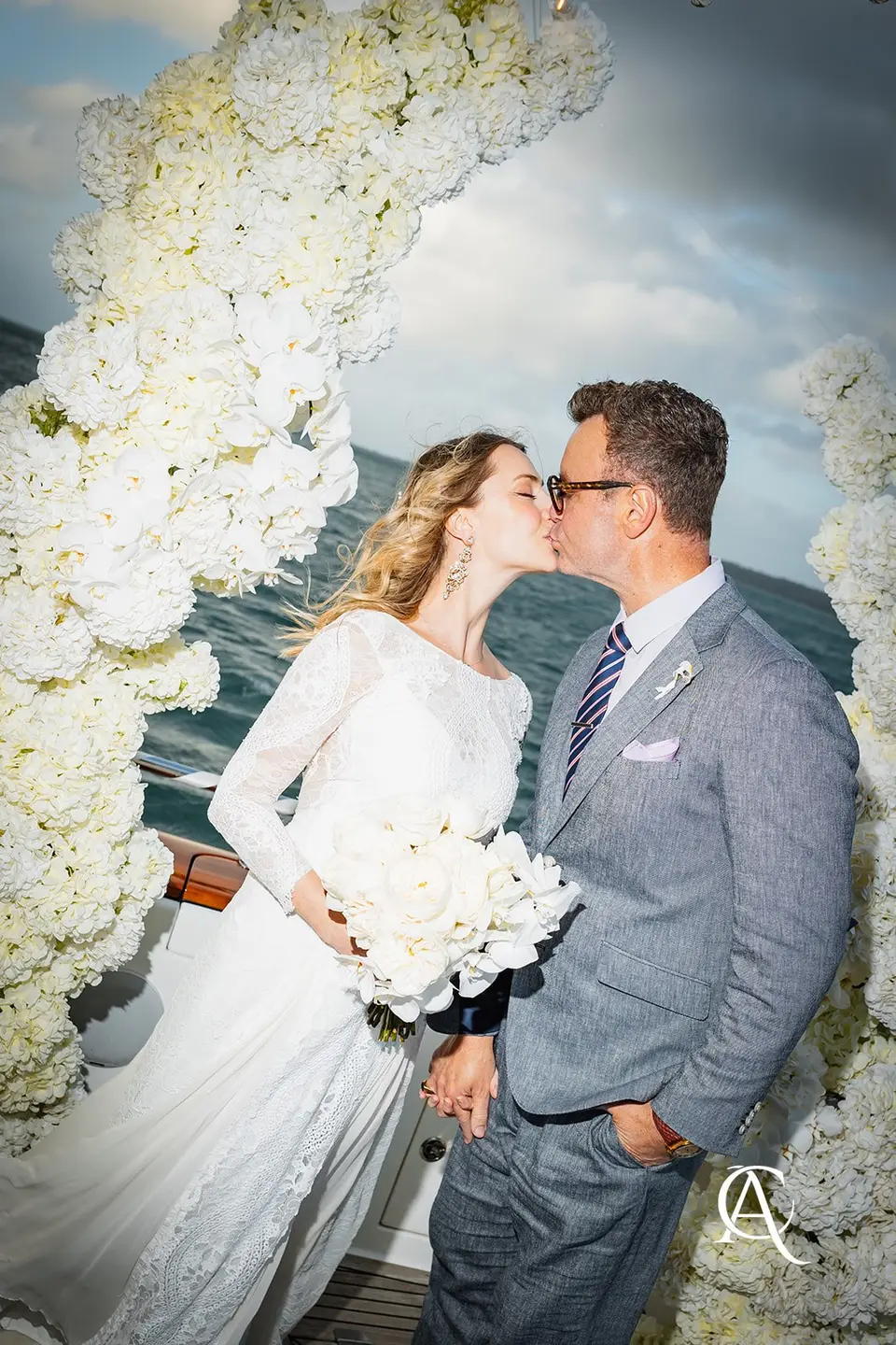 Couple kissing beside a tall column of white hydrangea and phalaenopsis orchids, the bride holding a white bouquet