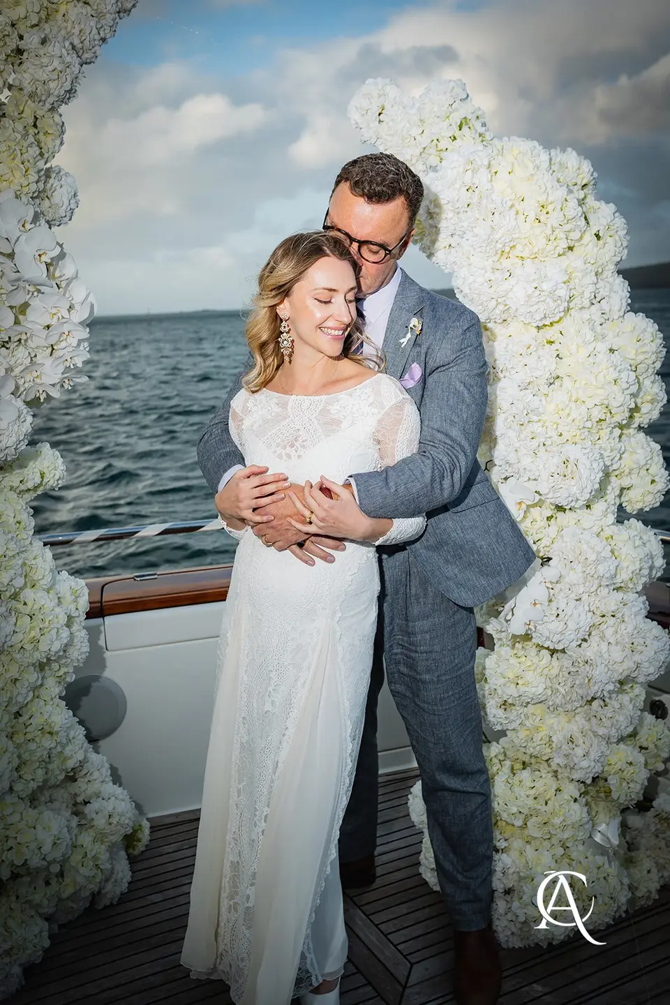 Groom holding the bride from behind between white hydrangea columns on a yacht, with the harbour and cloud behind