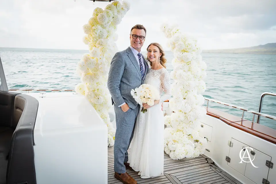 Couple smiling on a teak yacht deck between two white hydrangea columns, with the harbour and Rangitoto behind