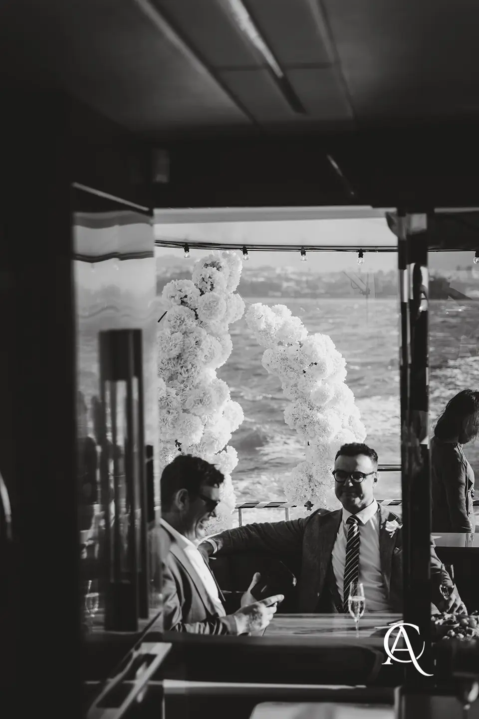 Black and white photo of guests talking at a table inside a yacht, with the white hydrangea columns and the harbour seen through the doorway