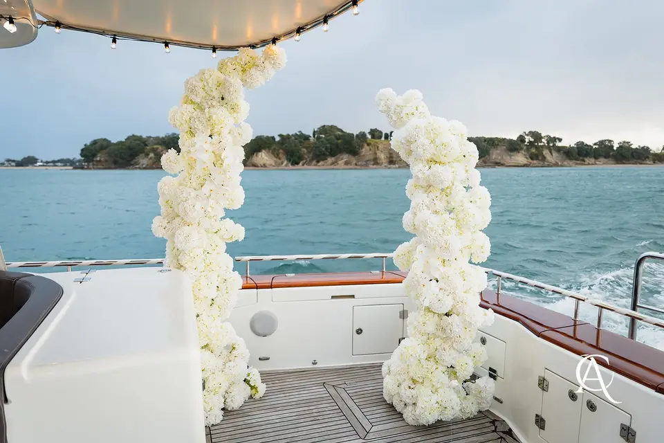 Two white hydrangea columns on an empty yacht deck under a canopy of festoon lights, with a bush-covered coastline behind