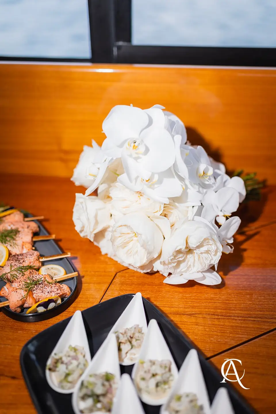 Bouquet of white garden roses and phalaenopsis orchids resting on a teak table beside platters of canapés