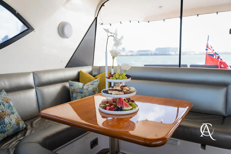 White rose and orchid bud vases with a tiered dessert stand on a teak table in a yacht lounge, the harbour behind